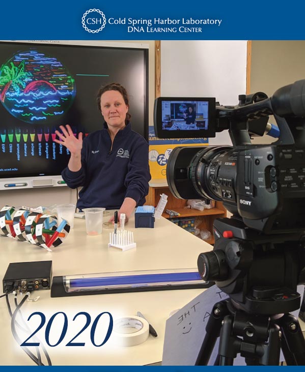 Annual report cover with a DNALC educator in front of a smartboard with fluorescent art on a petri dish and fluoescent liquids in test tubes, who is waving at a large studio camera during filming of a virtual laboratory demonstration